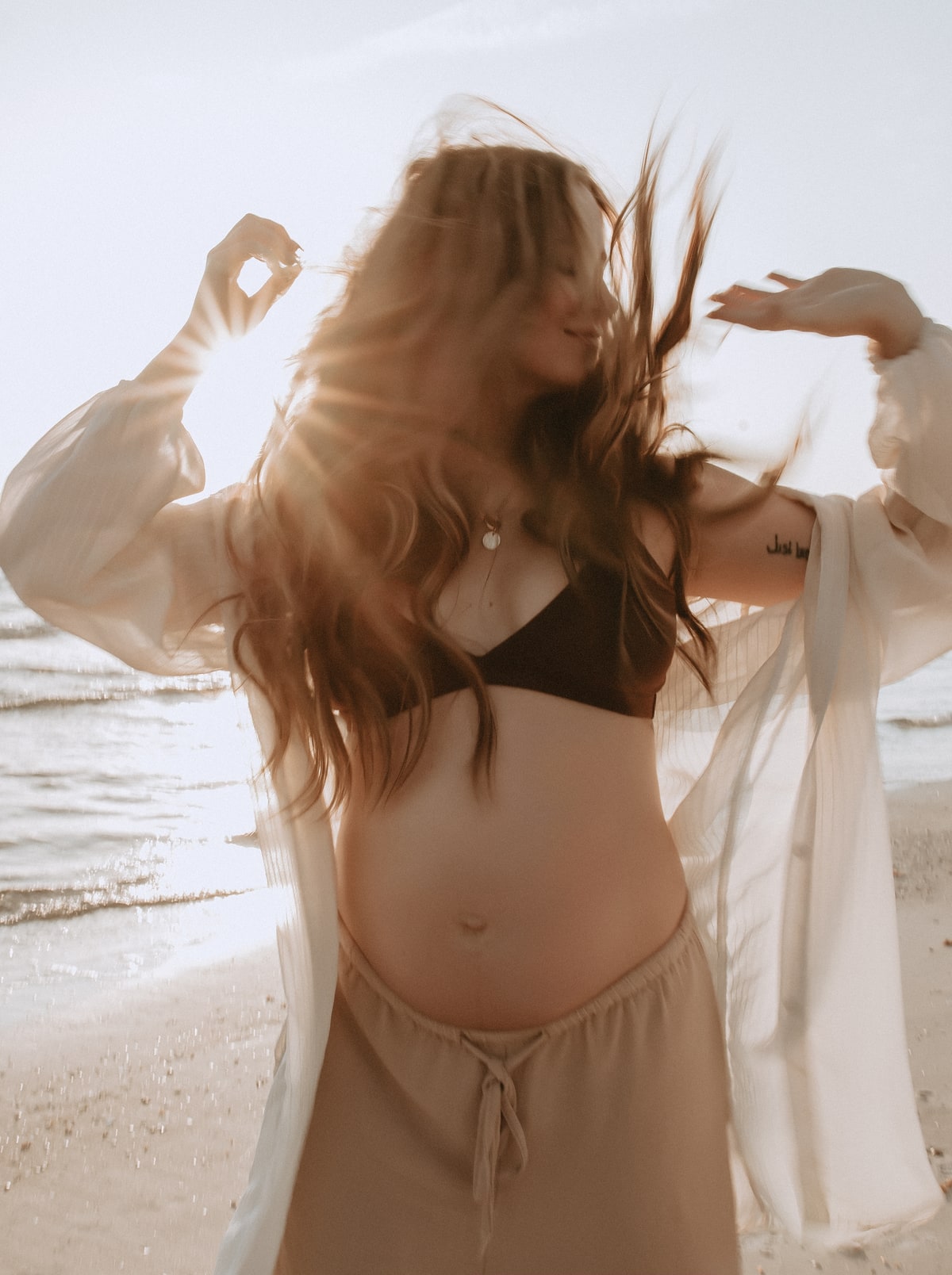 Madison, pregnant, smiling on the beach at sunset, her hair blowing, in a sheer white cover-up.